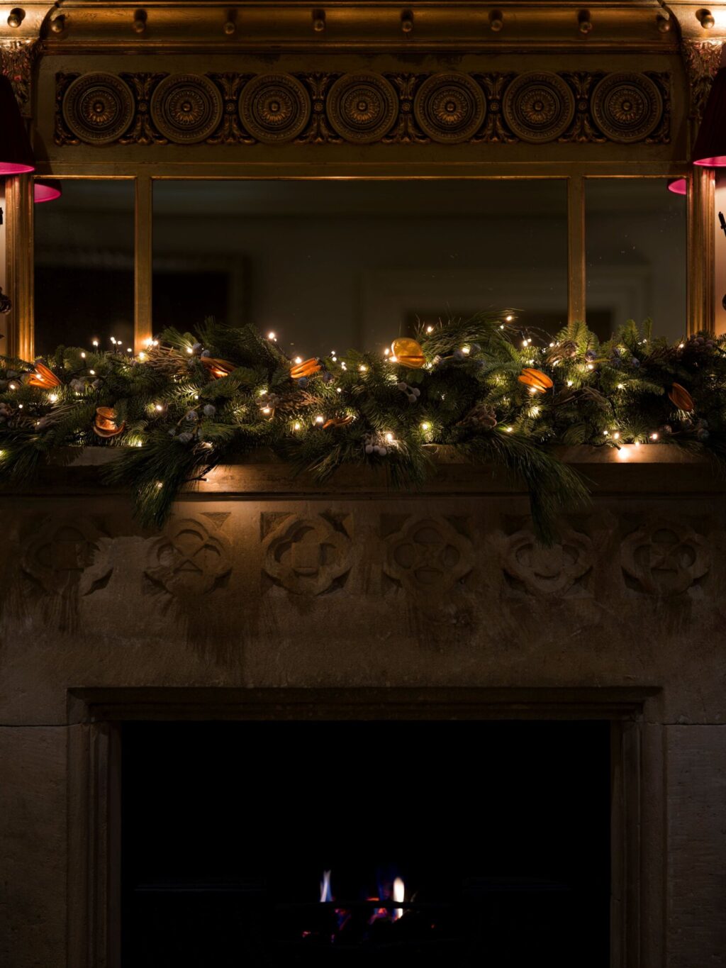 A softly glowing stone fireplace at Hazlitt’s decorated with a lush Christmas garland of greenery, dried oranges, and warm fairy lights beneath a gilded mirror.