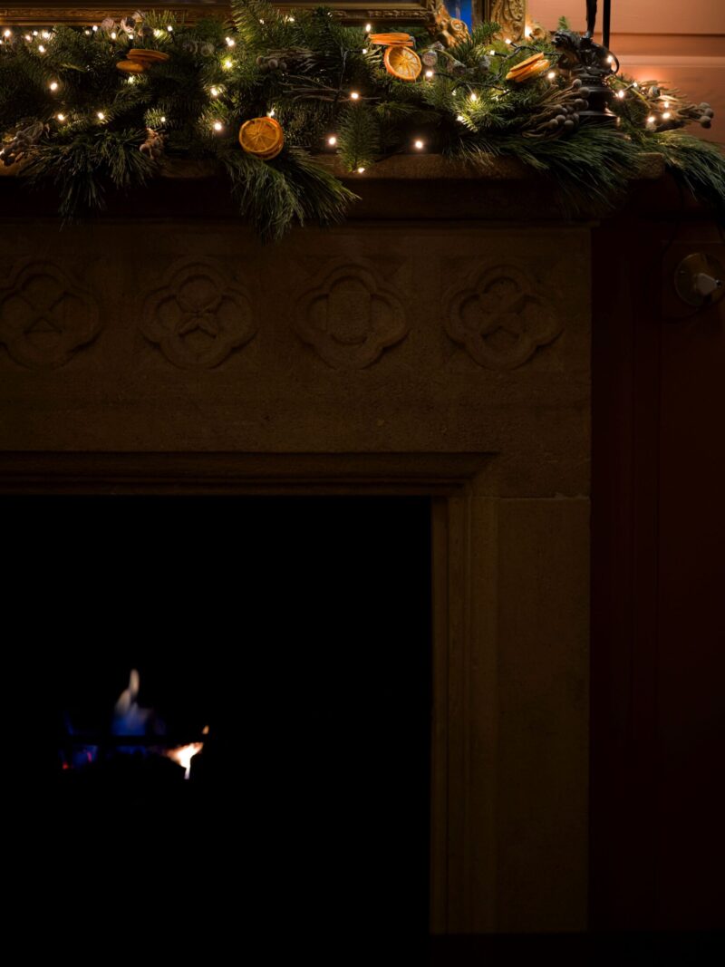 A stone fireplace at Hazlitt’s decorated with a Christmas garland of greenery, dried oranges, and warm fairy lights, with a small flame glowing in the dark hearth below.