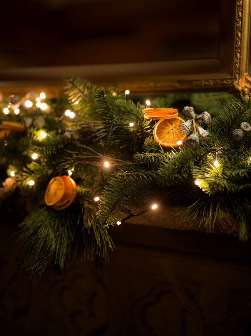 Close-up of a Christmas garland with pine branches, dried orange slices, berries, and warm fairy lights on a stone mantel at Hazlitt’s.