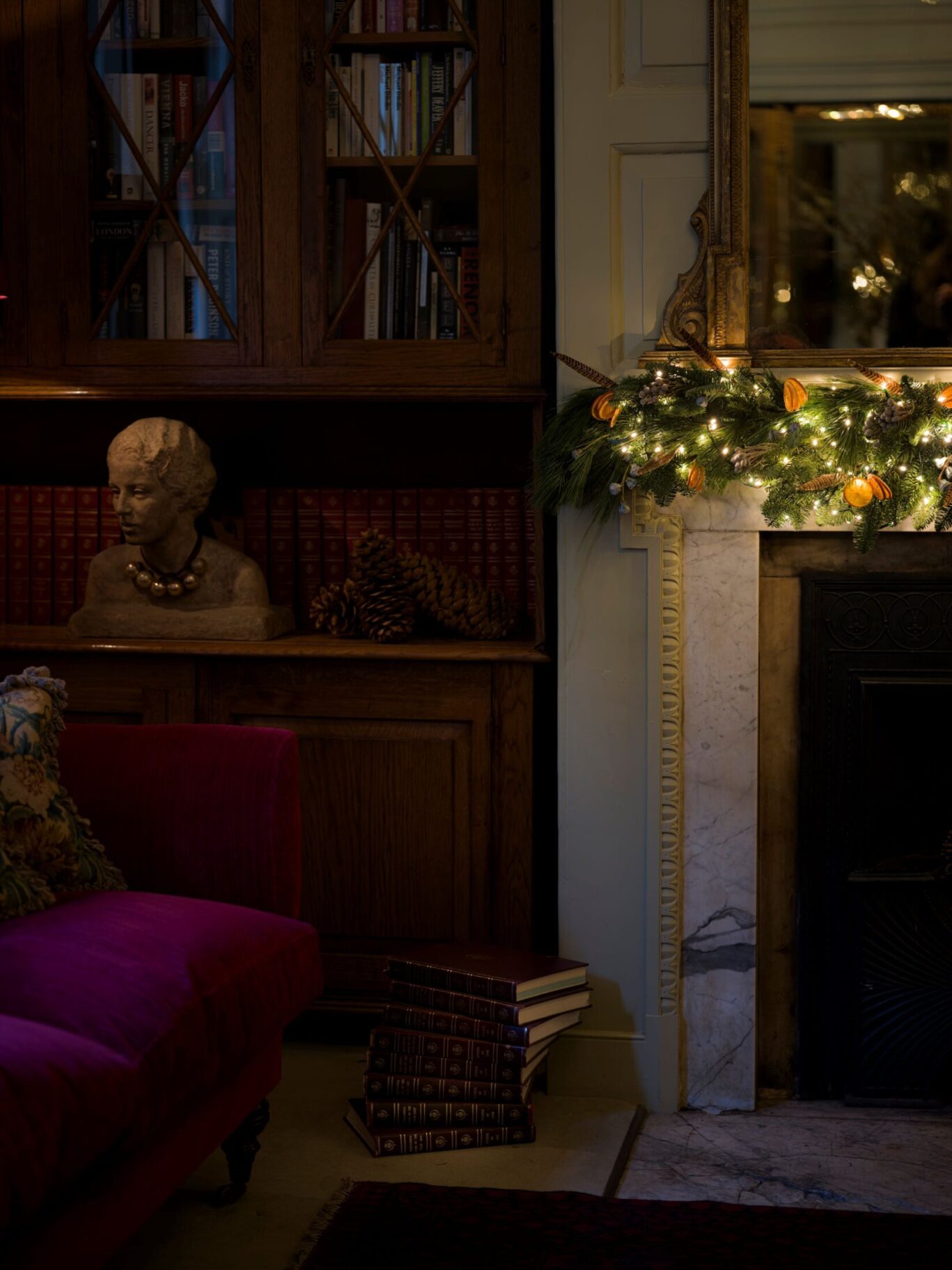 A festive garland with lights and dried oranges decorates a marble fireplace beside a wooden bookcase and red velvet sofa at Hazlitt’s.