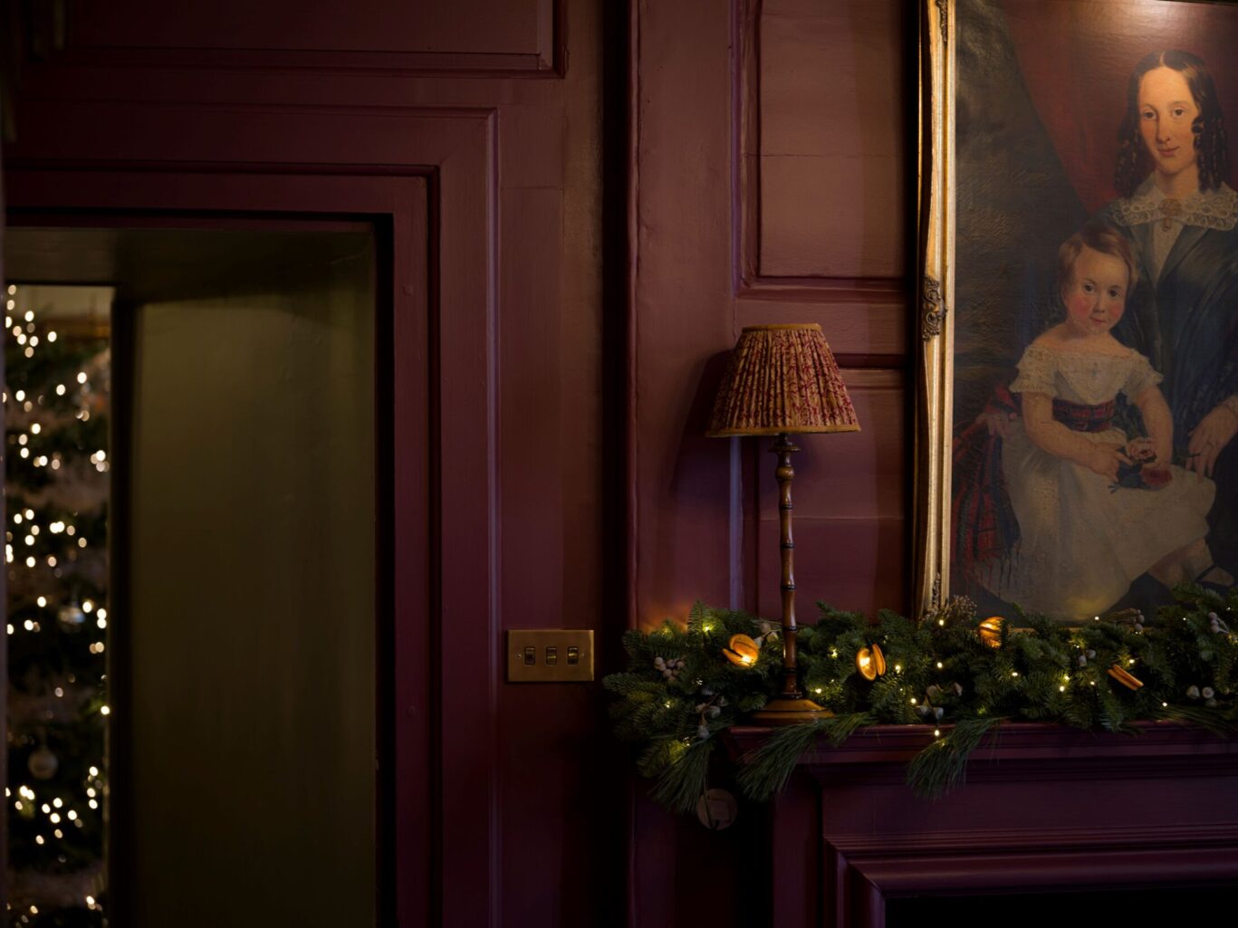 A mantel in the Sitting Room at Hazlitt’s decorated with evergreen garland and warm lights beneath a large vintage portrait, with a glimpse of a Christmas tree through a doorway.