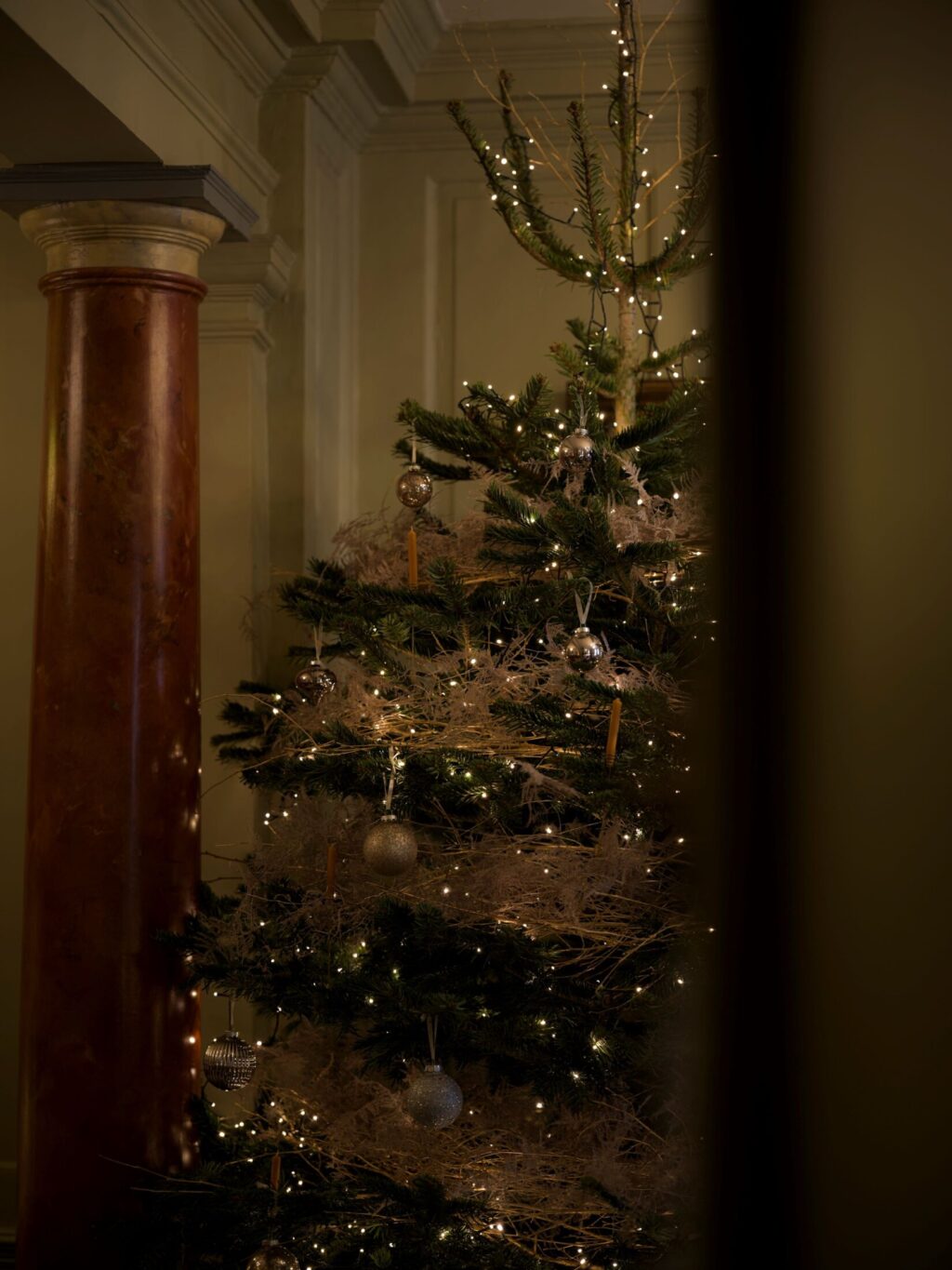 A softly lit Christmas tree decorated with warm lights and silver ornaments stands beside a marbled column at Hazlitt’s.